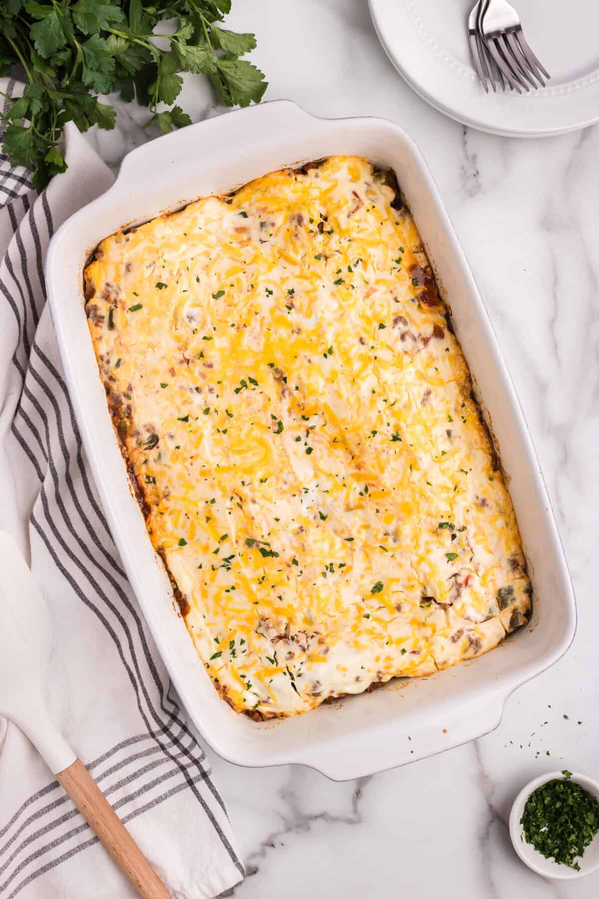 A fully baked casserole with a golden, bubbly cheese topping, garnished with finely chopped herbs. The dish is placed on a marble countertop with a striped kitchen towel, a spatula, and fresh parsley nearby.
