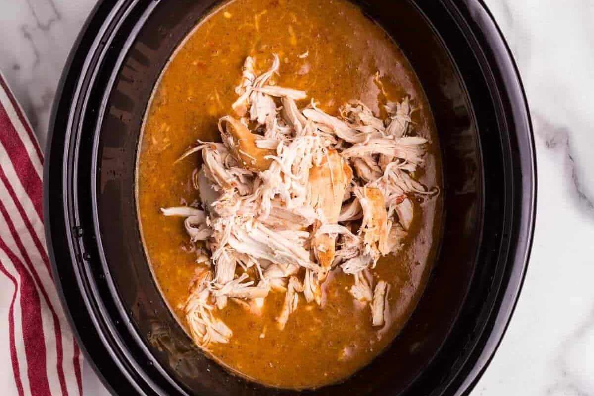 A black slow cooker filled with rich, golden King Ranch Chicken Soup, with freshly shredded chicken added back into the broth. A red-striped kitchen towel rests on the white marble countertop beside the slow cooker.