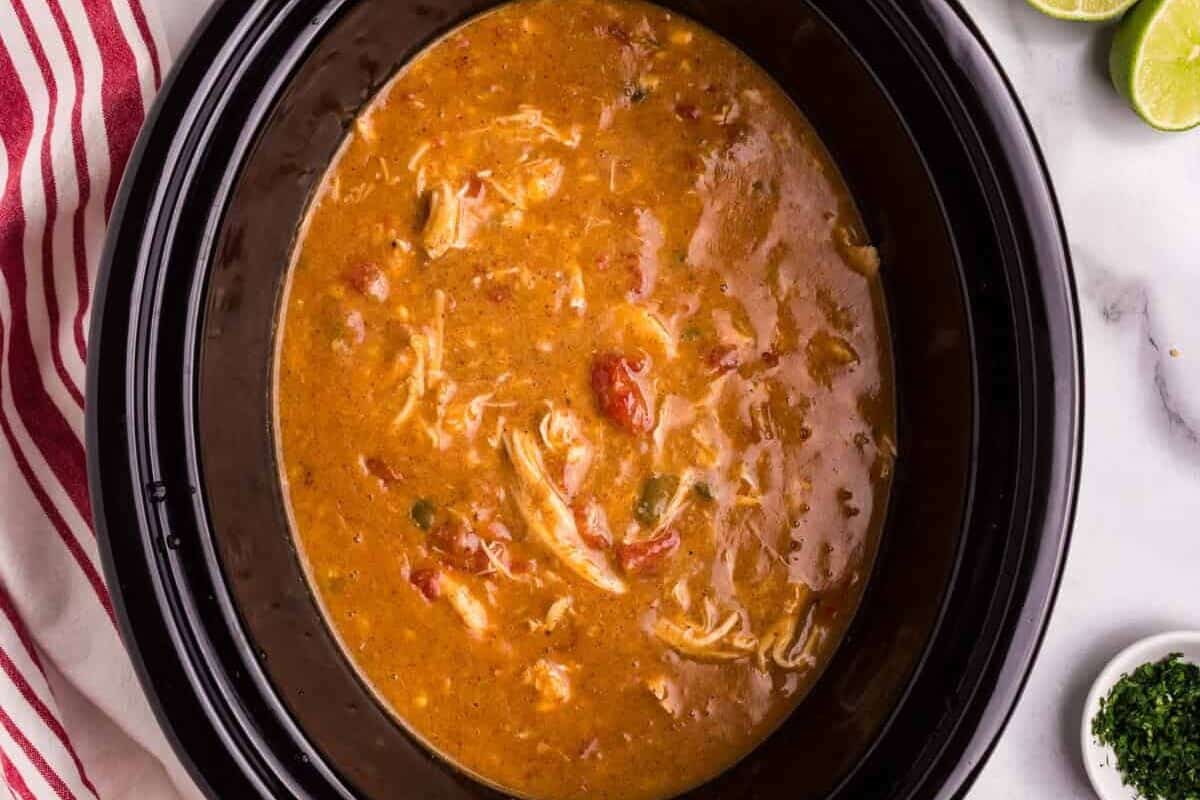 A black slow cooker filled with freshly made King Ranch Chicken Soup, featuring a rich, golden broth with shredded chicken and diced tomatoes. Surrounding the slow cooker are garnishes including sliced jalapeños, fresh cilantro, lime halves, tortilla chips, and shredded cheese. A red-striped kitchen towel rests on the white marble countertop.