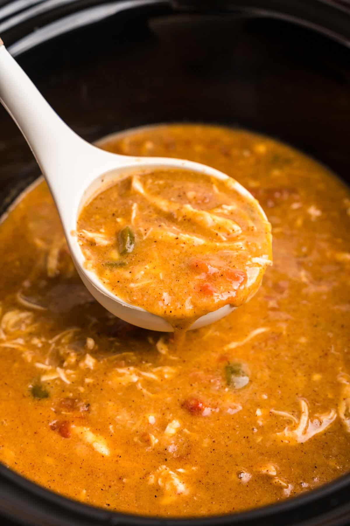 A close-up of a white ladle scooping a serving of King Ranch Chicken Soup from a slow cooker. The soup has a rich, golden-brown broth with shredded chicken, diced tomatoes, green peppers, and a blend of spices. The ladle is slightly dripping, showcasing the creamy and hearty texture of the dish.





