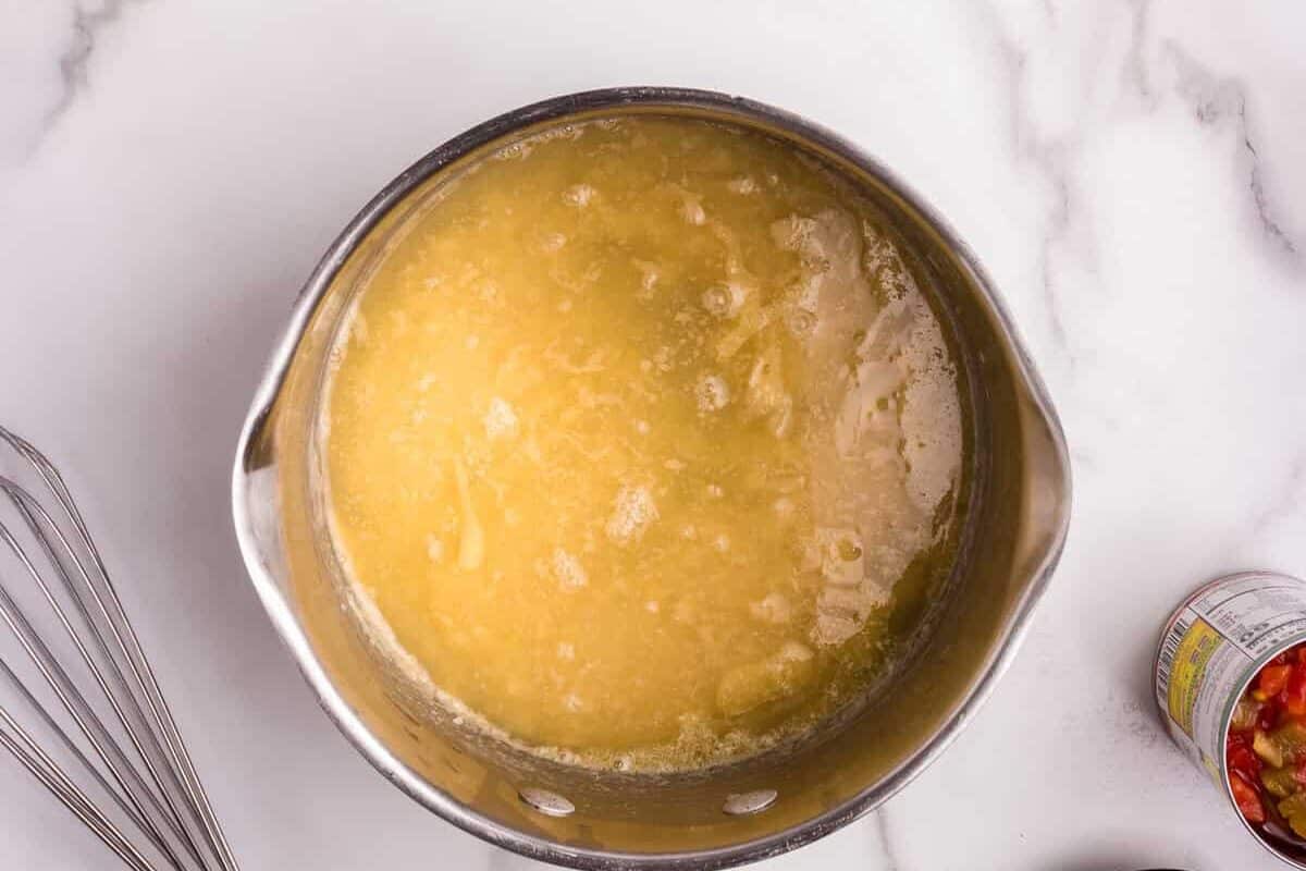 A stainless steel saucepan with melted butter and flour bubbling together, forming the base for a roux. Surrounding the pan are small bowls of spices, a measuring cup filled with chicken broth, chopped jalapeños, and a can of diced tomatoes with green chilies. A wooden-handled whisk rests nearby, ready for stirring.