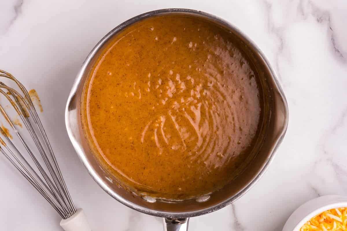 A stainless steel saucepan filled with a rich, golden-brown spiced sauce, whisked to a smooth consistency. Surrounding the saucepan are raw chicken breasts on a white plate, a can of diced tomatoes with green chilies, a small bowl of chopped jalapeños, a whisk with sauce residue, and a bowl of shredded cheese, all set on a clean, white marble surface, ready for the next steps in the cooking process.