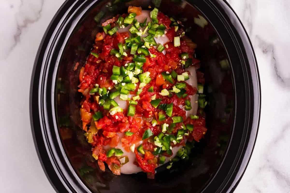 A black slow cooker containing raw chicken breasts topped with diced tomatoes and chopped jalapeños, ready for cooking. Surrounding the slow cooker are a saucepan with a rich golden-brown spiced sauce, a bowl of shredded cheese, and a small empty black dish on a white marble countertop.