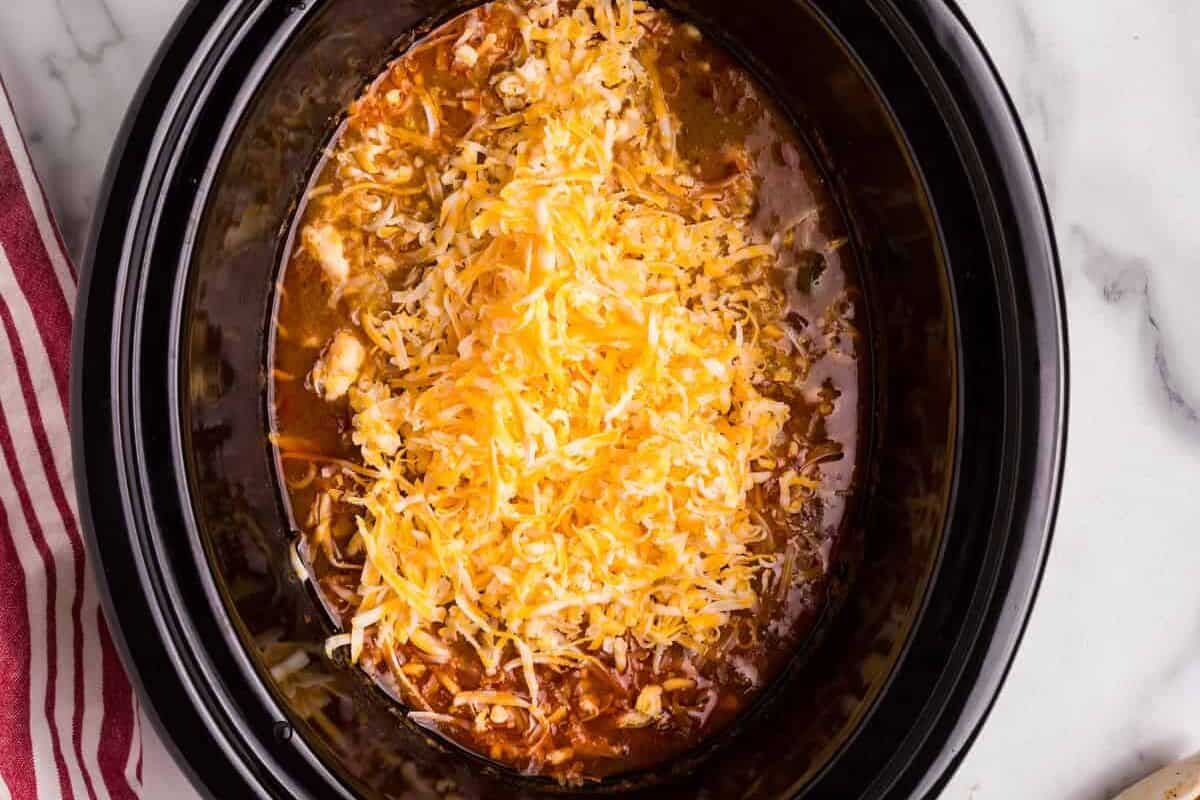 A black slow cooker filled with a rich, simmering King Ranch Chicken Soup, topped with a generous amount of shredded cheddar and Monterey Jack cheese. Cooked chicken breasts are set aside on a white plate, ready to be shredded and added back to the dish. Tongs and a red-striped kitchen towel rest on a white marble countertop.