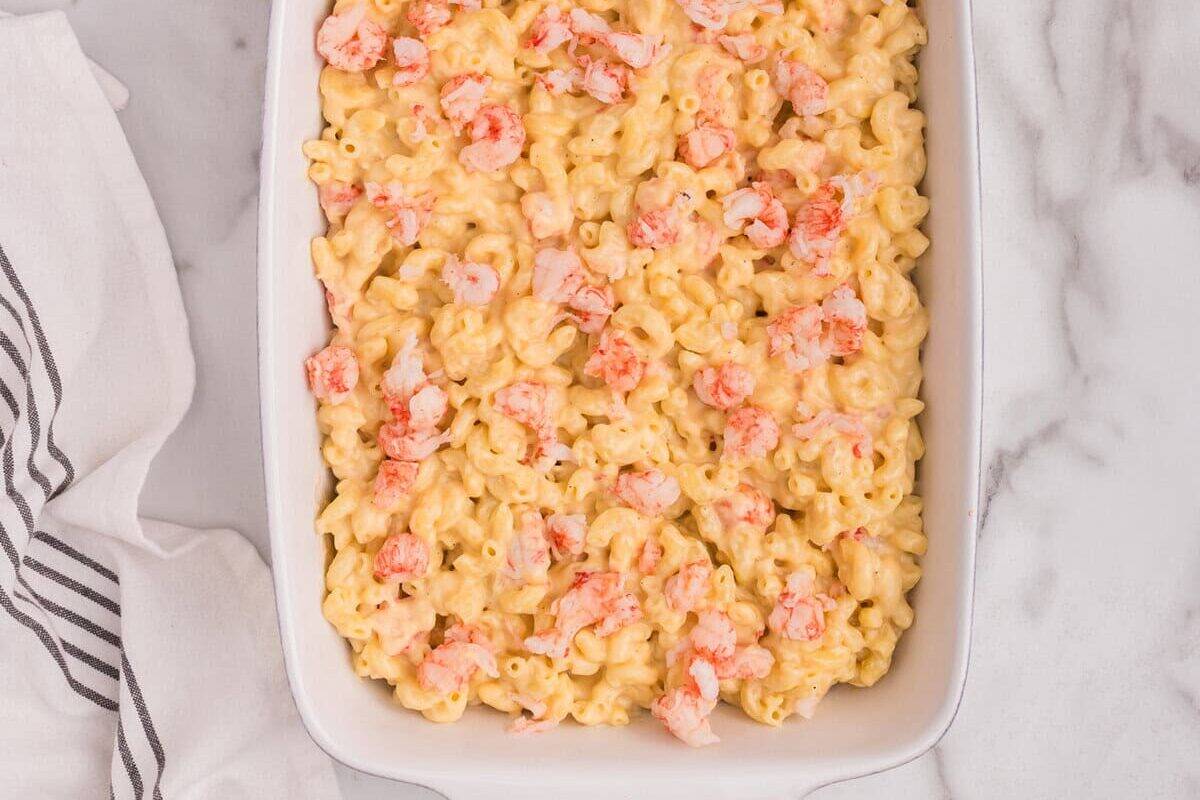 Baking dish filled with macaroni and cheese mixed with lobster, topped with extra lobster pieces, with a bowl of breadcrumbs nearby.