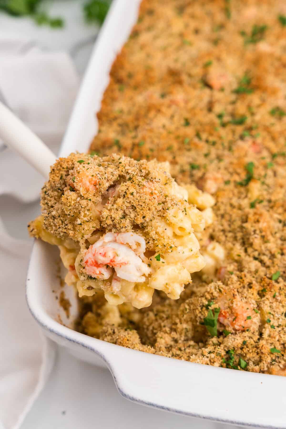 Close-up of a serving spoon lifting a portion of lobster mac and cheese from the baking dish, showing lobster chunks and golden breadcrumb topping.

