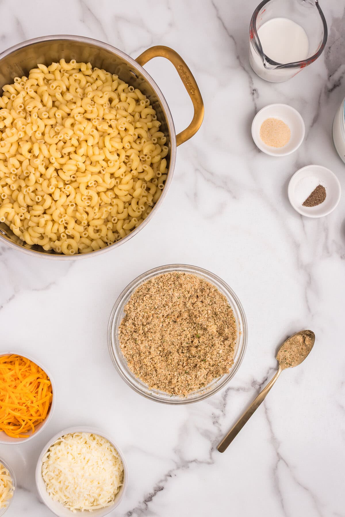 Overhead view of macaroni in a pot, with the breadcrumb mixture now fully combined in the bowl, alongside shredded cheese, milk, and seasonings.