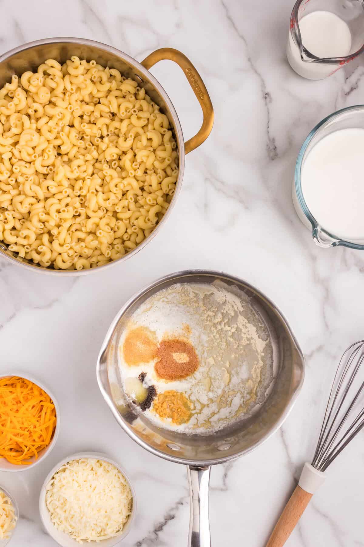 Overhead view of macaroni in a pot, with a saucepan containing melted butter and various seasonings before being mixed, surrounded by shredded cheeses and milk.