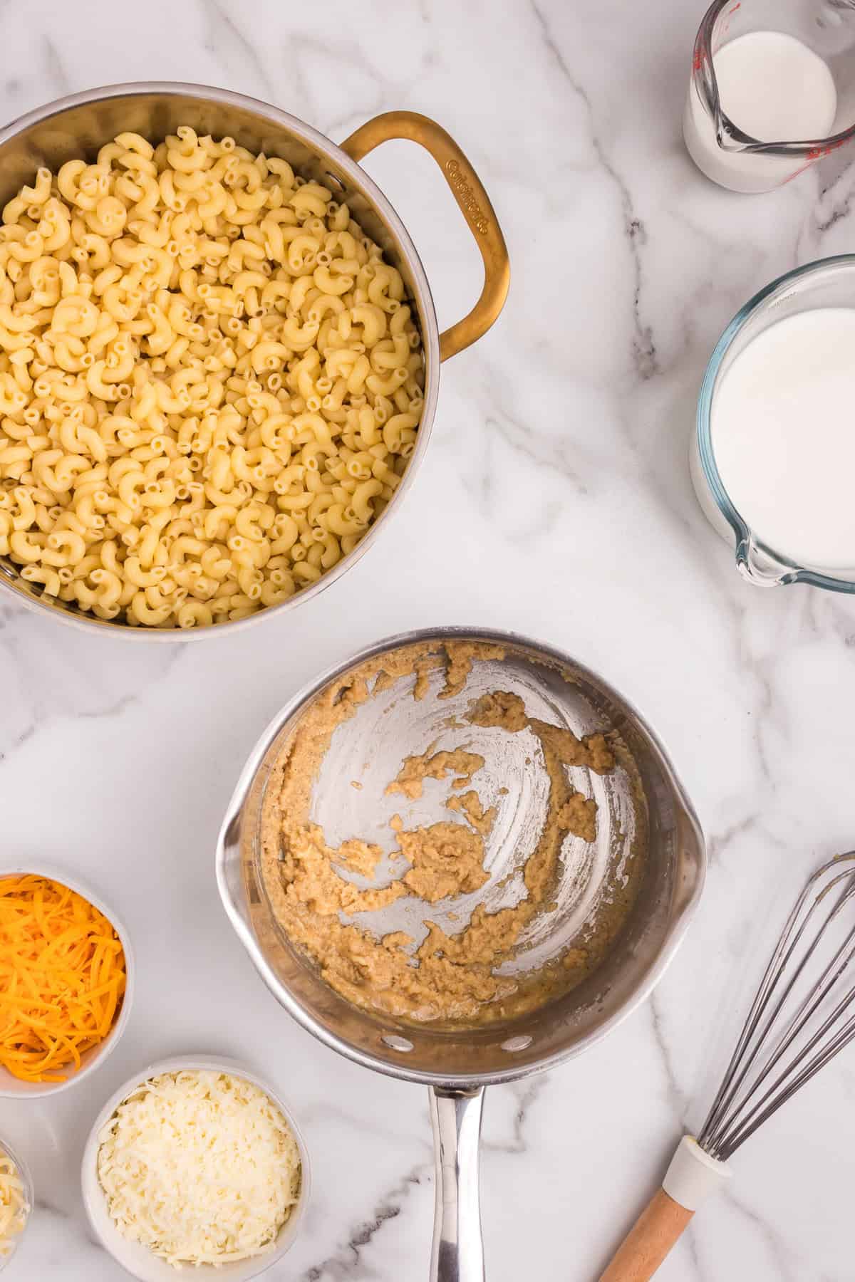 Overhead view of macaroni in a pot, with the saucepan now showing a thickened, combined seasoning paste, surrounded by shredded cheeses and milk.