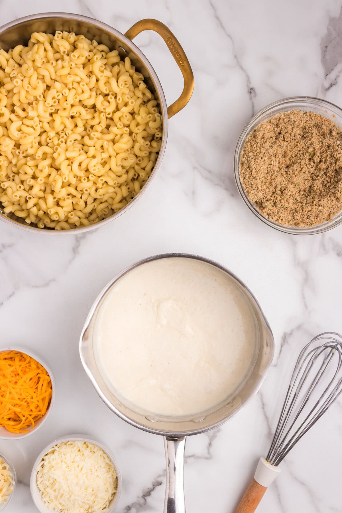 Overhead view of macaroni in a pot, with a saucepan containing a creamy white sauce, and the breadcrumb mixture in a bowl nearby.
