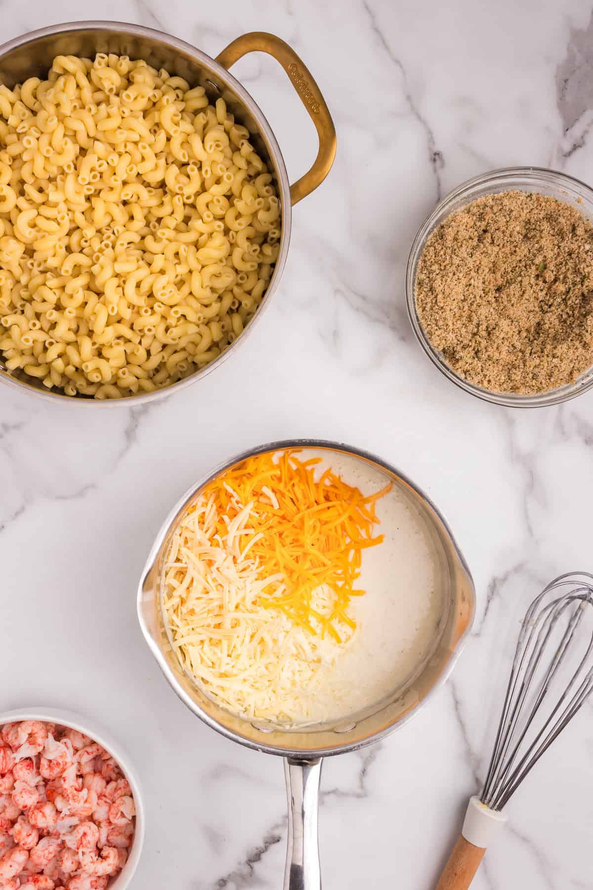 Overhead view of macaroni in a pot, with a saucepan of creamy sauce topped with shredded cheddar and mozzarella cheese, and a bowl of cooked lobster meat visible in the corner.