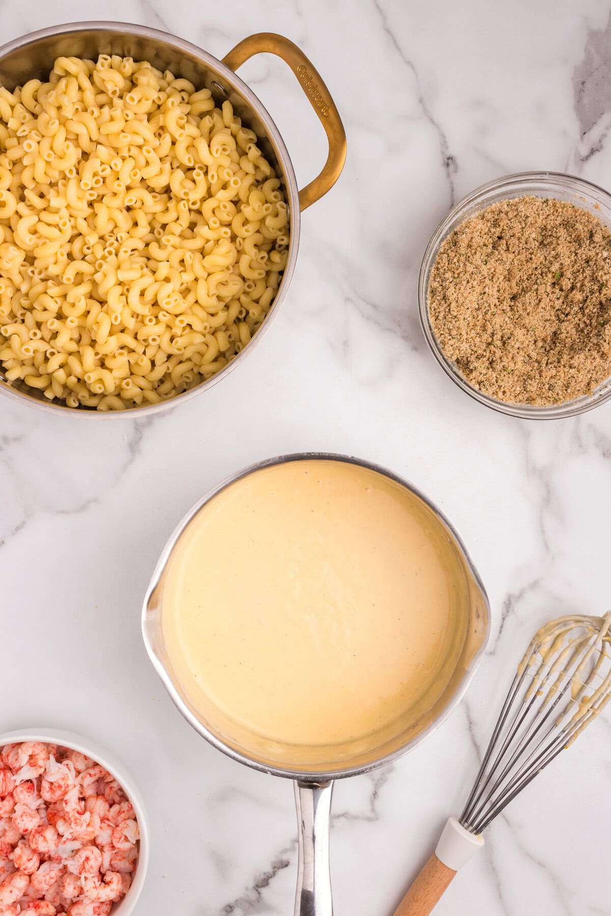 Overhead view of macaroni in a pot, with a saucepan of smooth, melted cheese sauce, alongside a bowl of lobster meat and breadcrumb mixture.