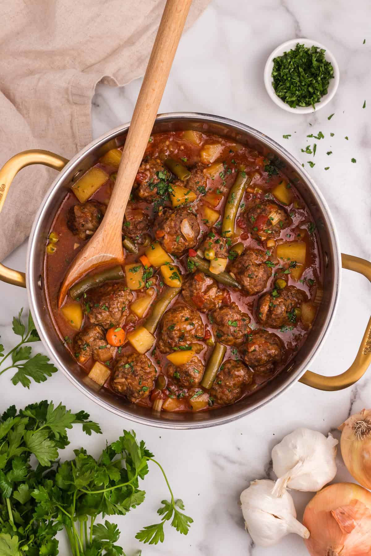 A pot of freshly cooked meatball stew with a wooden spoon resting inside. The stew is filled with juicy meatballs, diced potatoes, green beans, carrots, and peas in a rich tomato broth. Fresh parsley is sprinkled on top, and garnishes like garlic, onions, and parsley are arranged around the pot on a marble countertop.