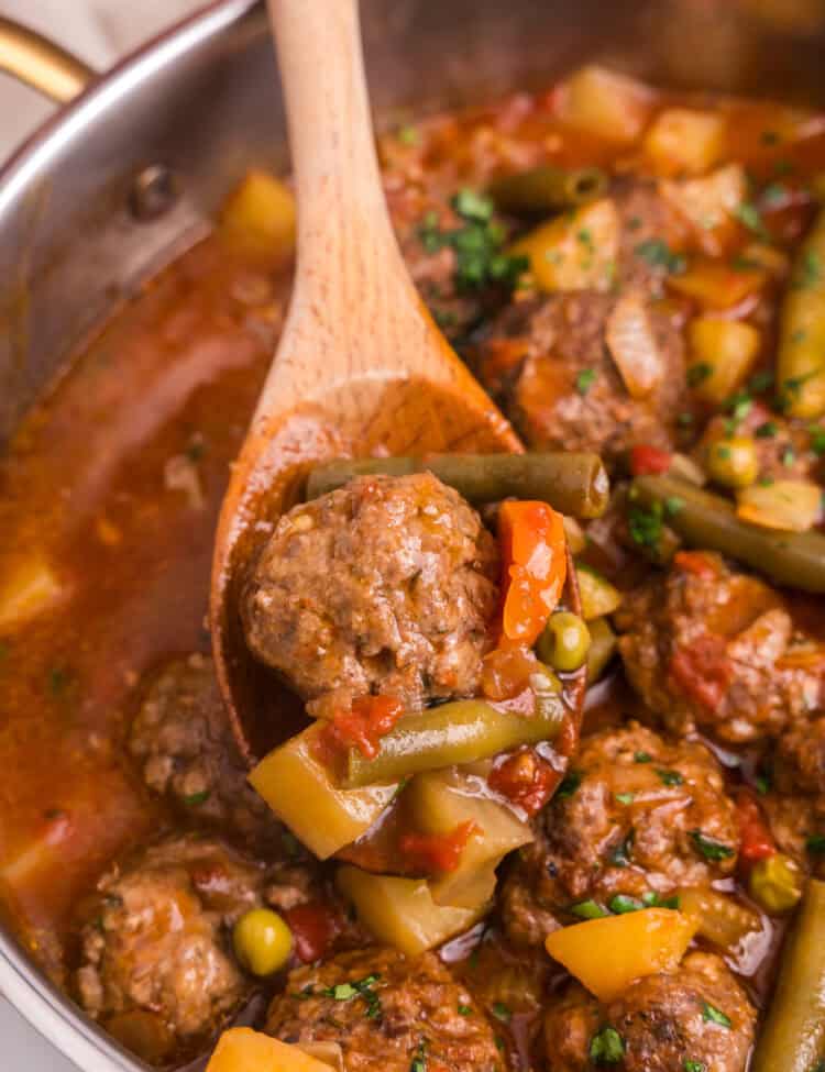 A wooden spoon lifts a serving of meatball stew from the pot, showcasing a tender meatball, green beans, peas, carrots, tomatoes, and potato chunks. The stew is rich and hearty, with visible chopped herbs adding color and freshness.
