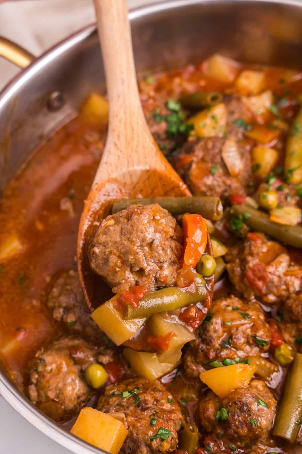 A wooden spoon lifts a serving of meatball stew from the pot, showcasing a tender meatball, green beans, peas, carrots, tomatoes, and potato chunks. The stew is rich and hearty, with visible chopped herbs adding color and freshness.

