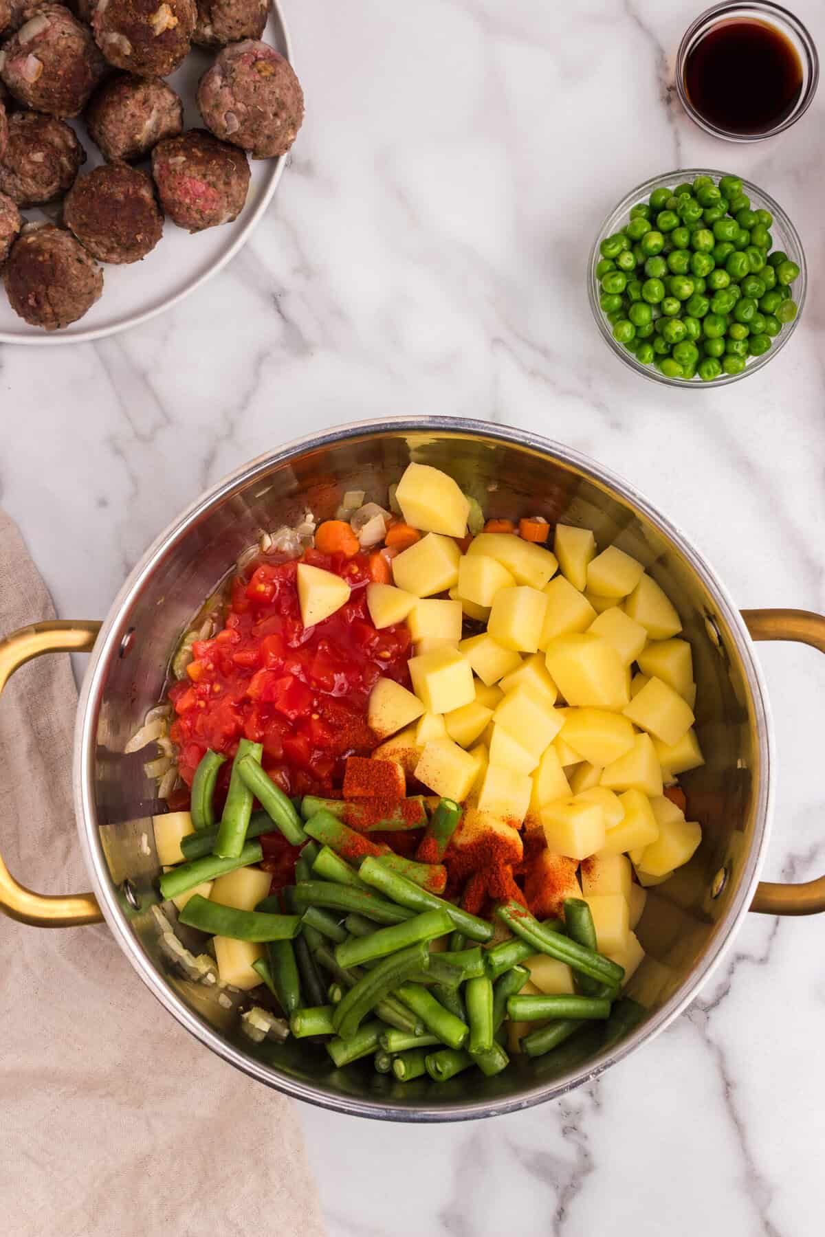 Diced potatoes, chopped tomatoes, green beans, and spices (like paprika) are added to the sautéed vegetables in the pot, ready to be mixed and simmered.