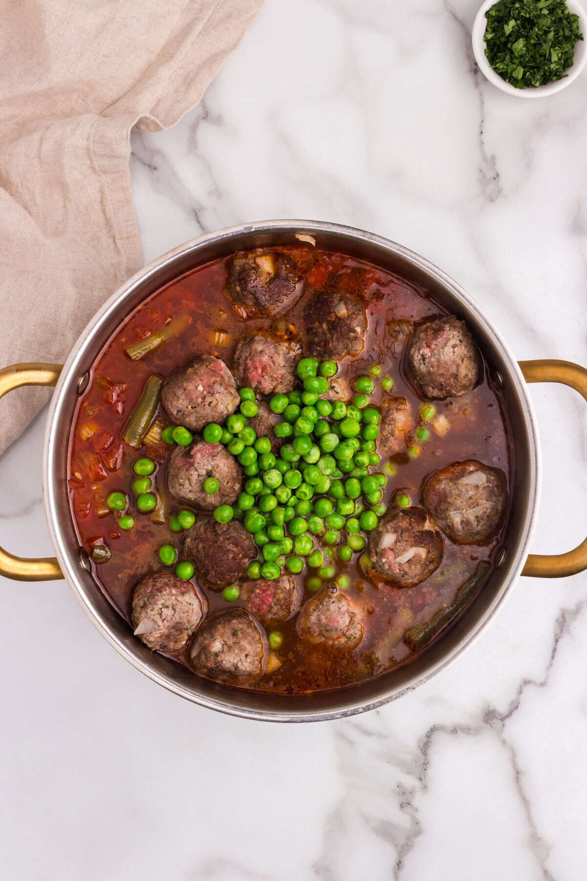 The browned meatballs are returned to the pot along with green peas, resting in a rich tomato-based broth. The stew is simmering to bring everything together.