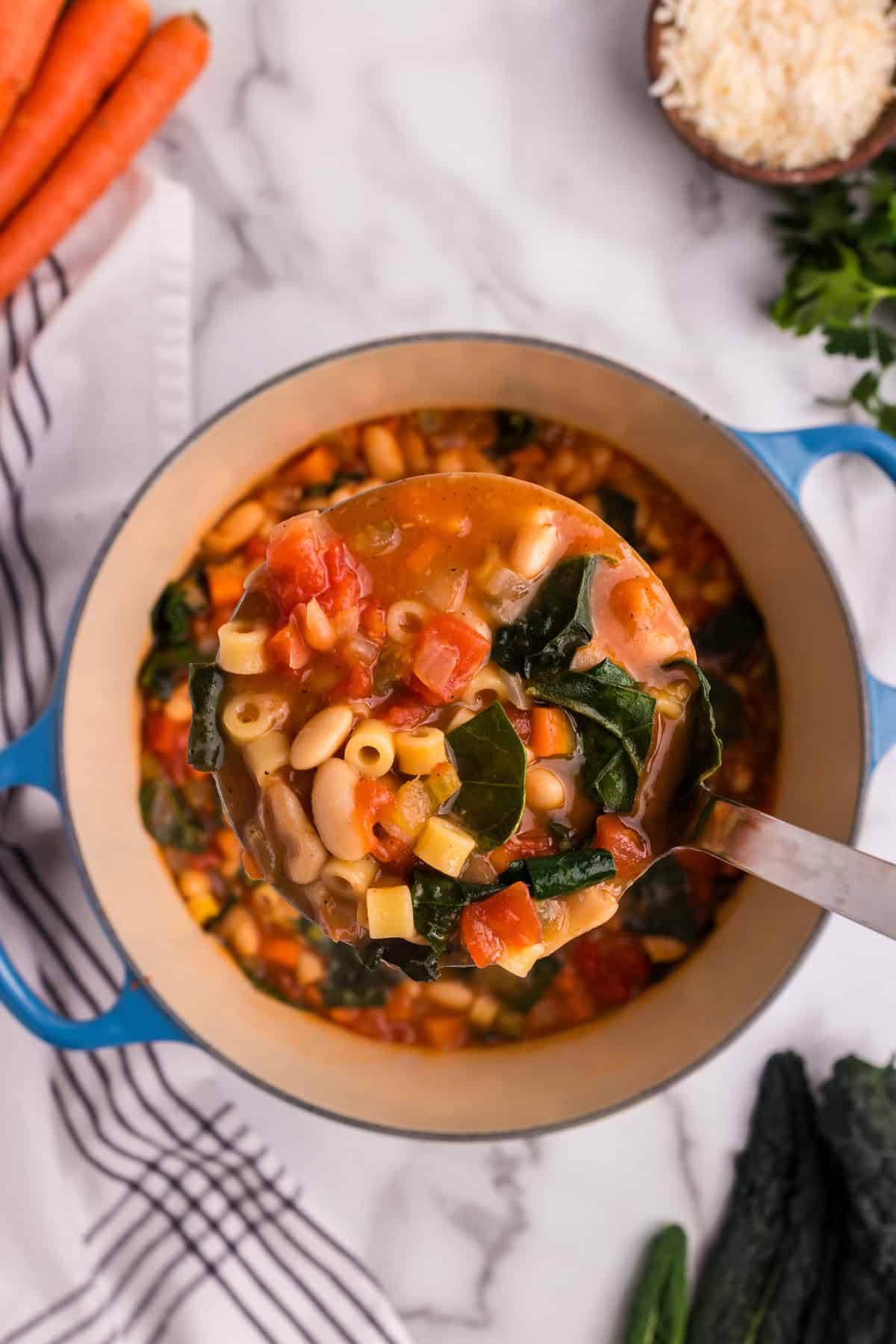 A ladle filled with hearty Pasta e Fagioli soup is lifted from a blue Dutch oven, showcasing ditalini pasta, cannellini beans, diced carrots, tomatoes, and kale in a rich tomato-based broth. The background features fresh carrots, a bowl of grated Parmesan cheese, and sprigs of parsley on a marble countertop, enhancing the rustic and homemade feel of the dish.