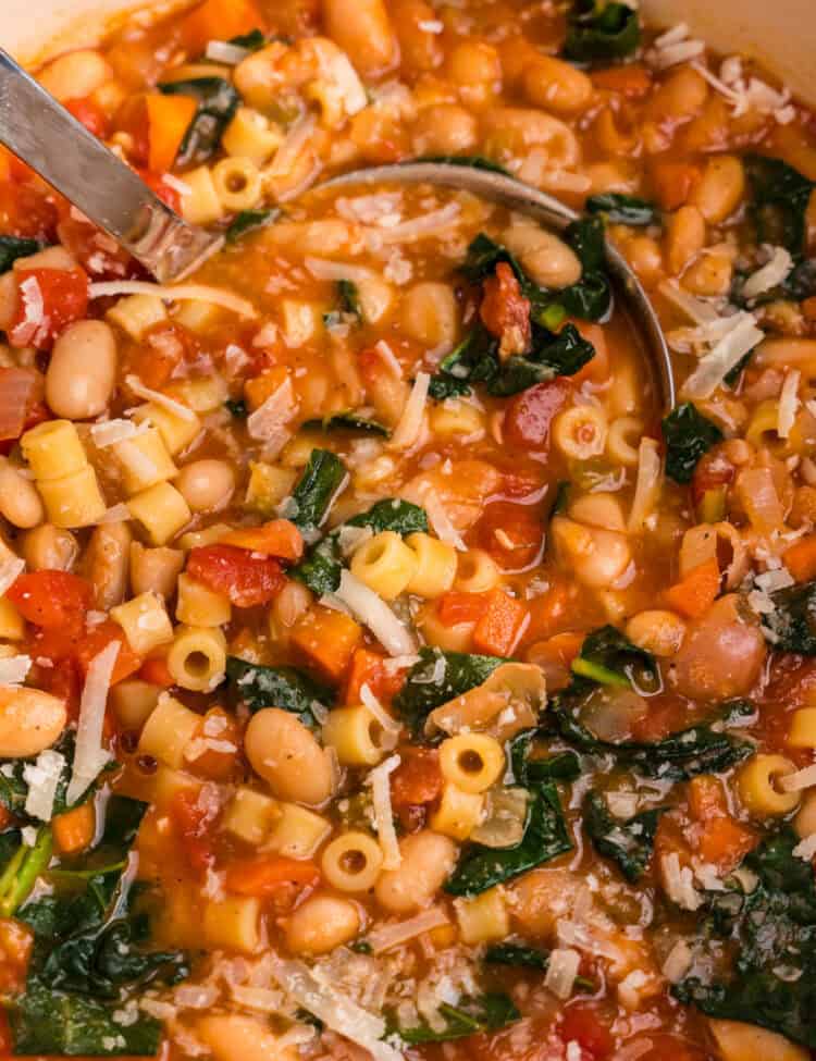 A close-up of a pot of Pasta e Fagioli soup, showing ditalini pasta, cannellini beans, diced carrots, tomatoes, and wilted kale in a rich, tomato-based broth. The soup is garnished with freshly grated Parmesan cheese, and a ladle is partially submerged, ready to serve. The colors and textures of the ingredients create a warm and inviting dish.