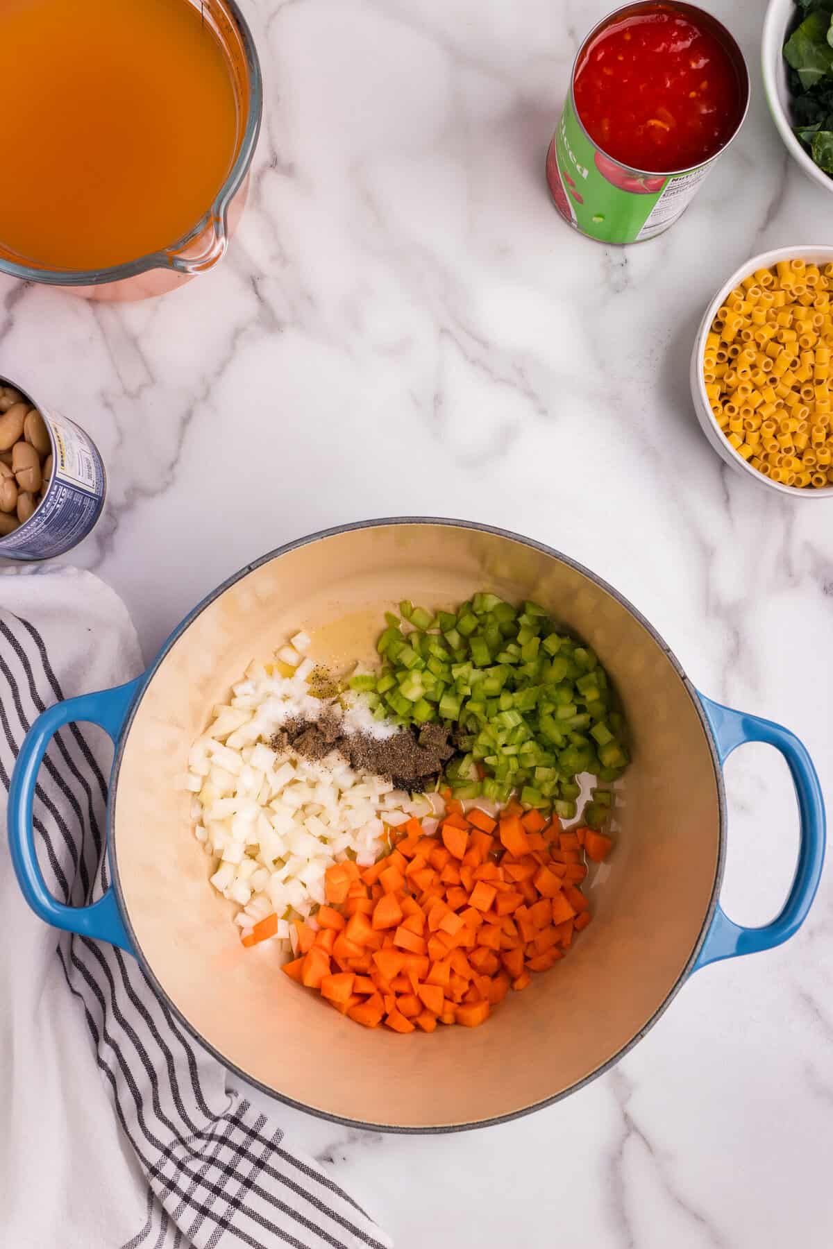 A blue Dutch oven sits on a marble countertop, filled with freshly chopped onions, celery, and carrots, along with salt and pepper, ready to be sautéed. Surrounding the pot are additional ingredients for Pasta e Fagioli, including vegetable broth, canned diced tomatoes, ditalini pasta, kale, and cannellini beans. A striped kitchen towel adds a cozy touch to the cooking scene.