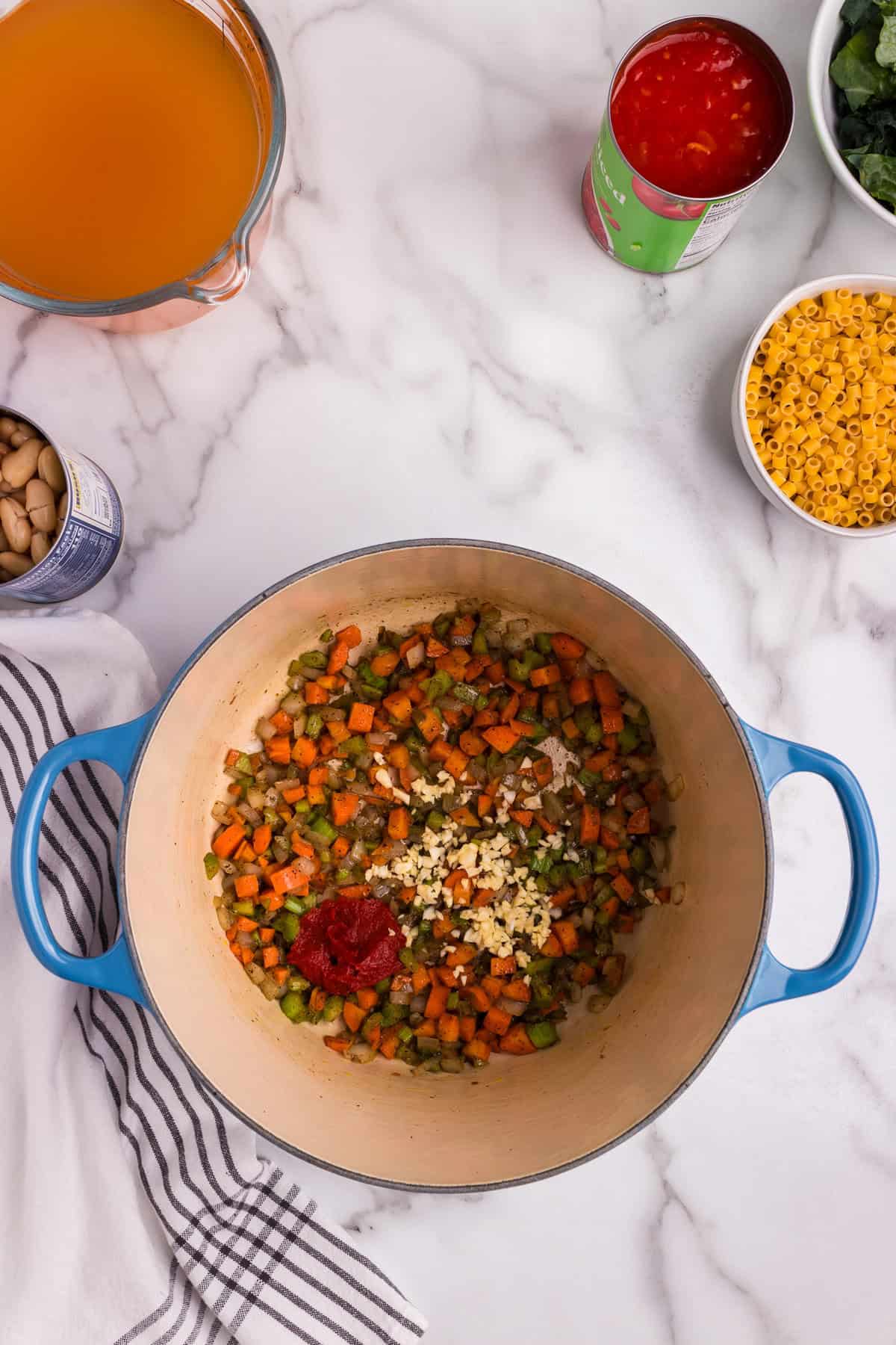 A blue Dutch oven sits on a marble countertop, containing sautéed diced carrots, celery, and onions seasoned with salt and pepper. Minced garlic and tomato paste have just been added to the mixture, ready to be stirred in. Surrounding the pot are ingredients such as vegetable broth, canned diced tomatoes, ditalini pasta, kale, and cannellini beans, arranged neatly for the next steps in the cooking process. A striped kitchen towel adds a cozy, homey touch to the scene.