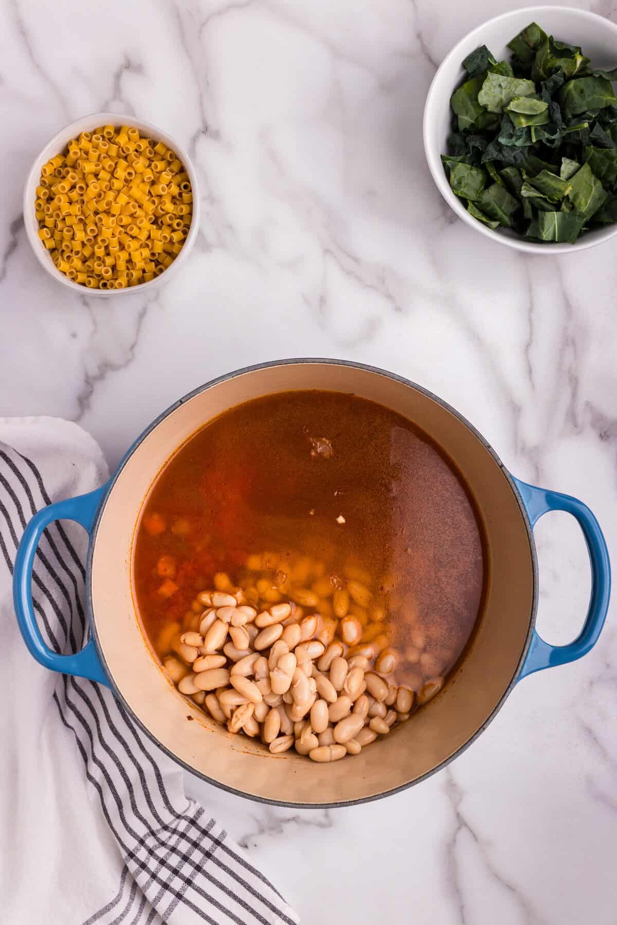 A blue Dutch oven filled with simmering vegetable broth and cannellini beans sits on a marble countertop. The broth has a rich, warm color, and the beans are freshly added. Nearby, a bowl of uncooked ditalini pasta and a bowl of chopped kale are ready to be incorporated into the soup. A striped kitchen towel is draped beside the pot, enhancing the cozy, homemade feel of the cooking process.