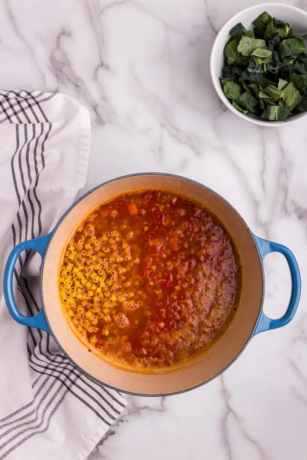 A blue Dutch oven filled with simmering Pasta e Fagioli soup sits on a marble countertop. Small ditalini pasta has just been added to the rich, tomato-based broth, alongside diced vegetables and beans. A bowl of chopped kale sits nearby, ready to be stirred into the soup. A striped kitchen towel is draped beside the pot, adding a cozy, homemade touch.