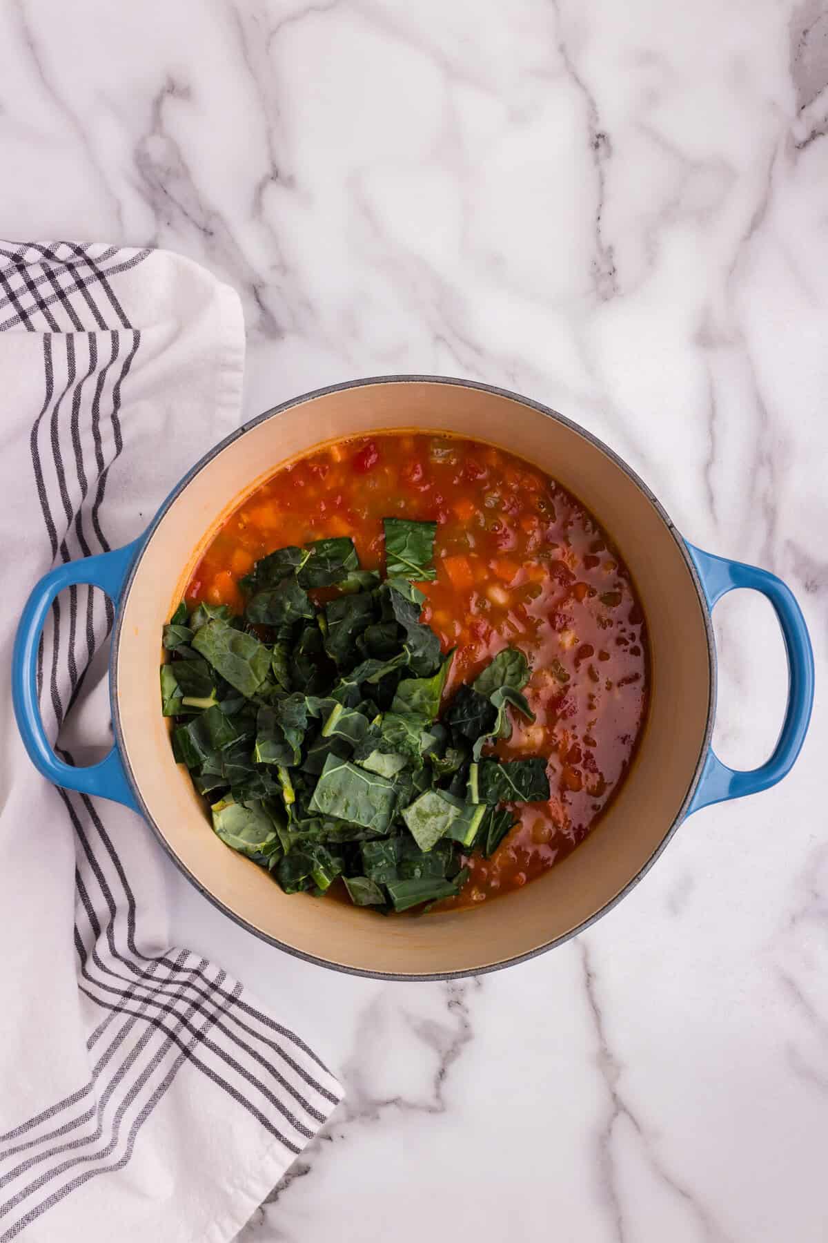 A blue Dutch oven filled with simmering Pasta e Fagioli soup sits on a marble countertop. The soup contains a rich, tomato-based broth with diced vegetables and beans. Freshly chopped kale has just been added on top, ready to be stirred in and wilt into the soup. A striped kitchen towel is draped beside the pot, adding a cozy, homemade touch.