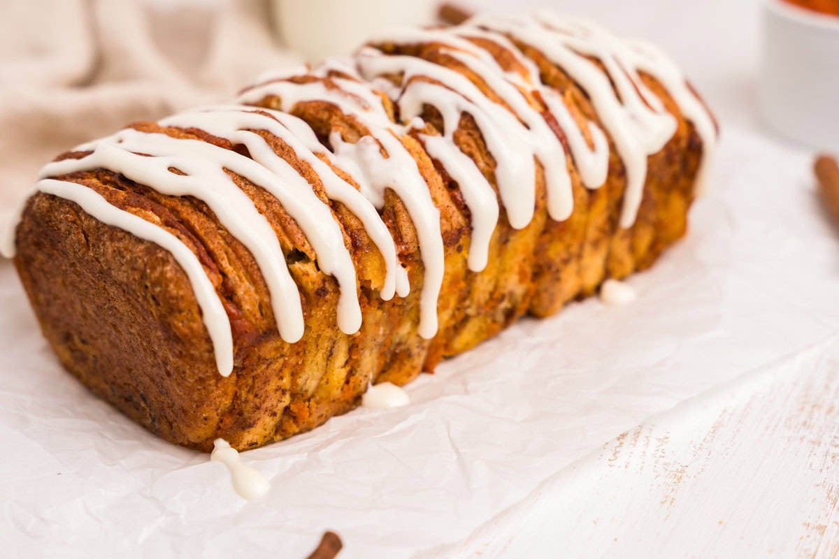 A loaf of pumpkin pull-apart bread topped with white icing drizzle, displayed on white parchment paper. The bread is golden brown with visible layers and a moist texture, giving it a rustic and appetizing appearance.