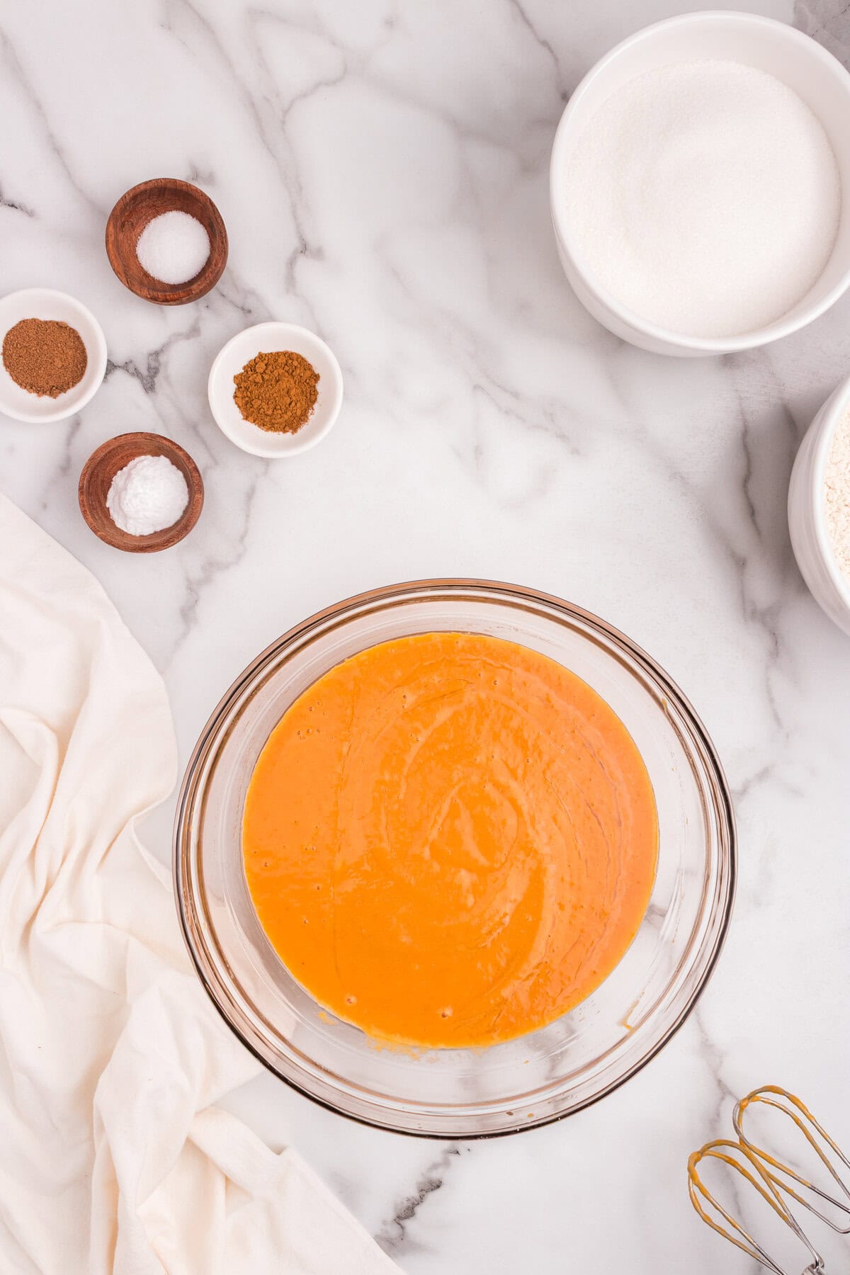 The ingredients in the glass bowl have been mixed into a smooth orange batter. The same small bowls of spices and sugar are visible around the bowl on the marble surface.