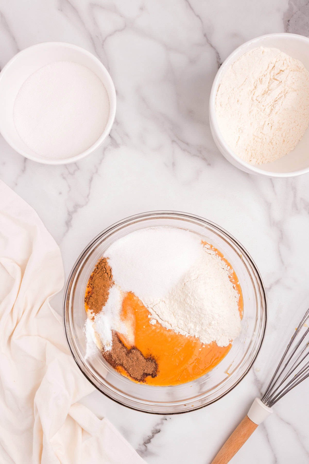 The batter in the bowl is now topped with flour, sugar, and spices that have just been added but not yet mixed in. The surrounding area still shows bowls of sugar and flour.