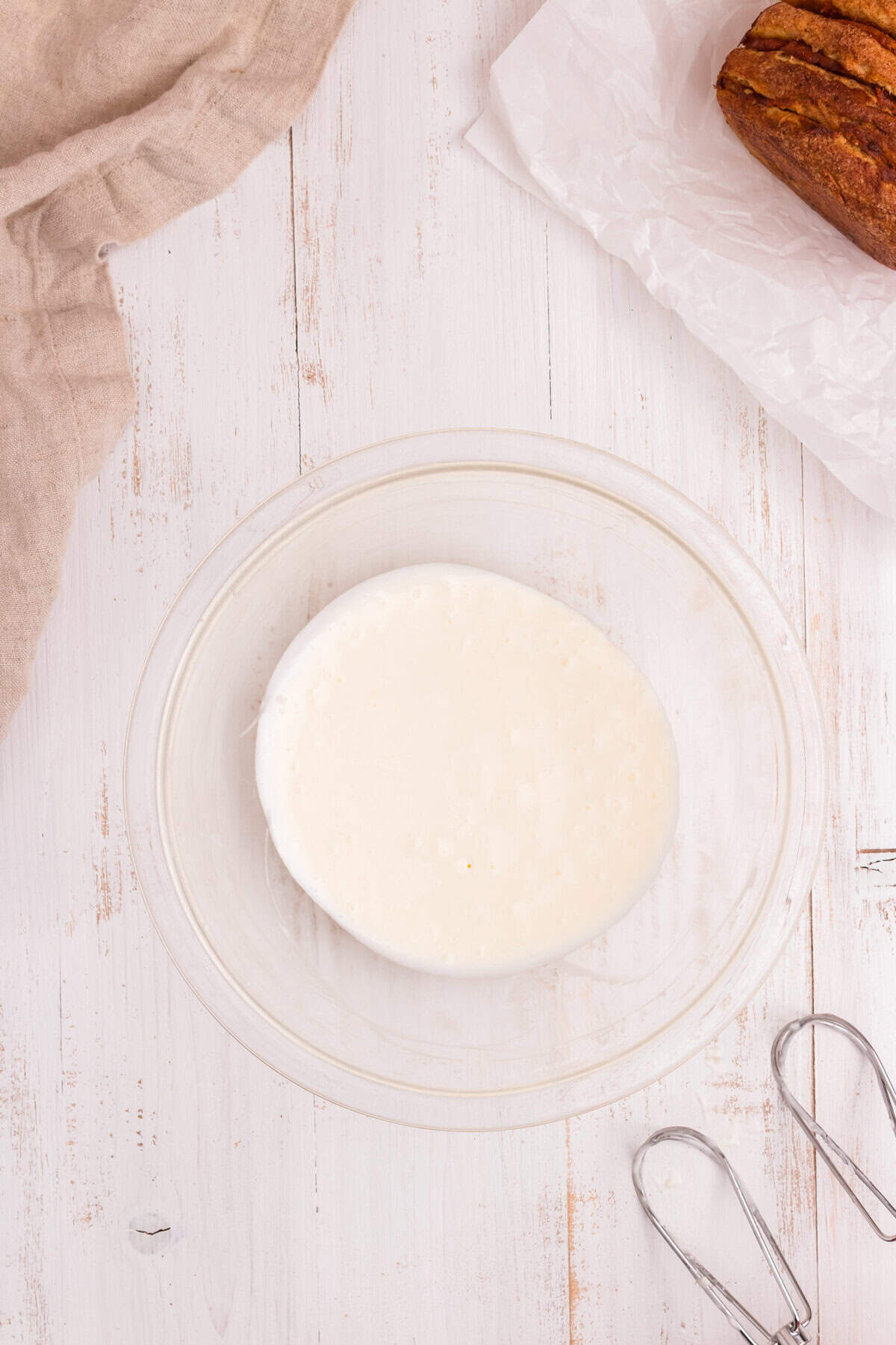 A glass mixing bowl filled with freshly made white icing glaze. An electric beater's whisk attachments rest beside the bowl, and part of the finished loaf is visible in the corner.