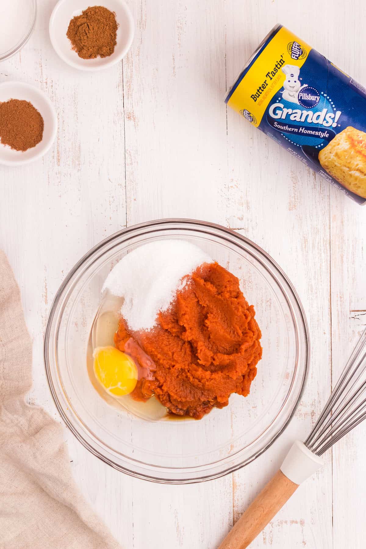 Ingredients for the pumpkin filling in a glass bowl, including canned pumpkin puree, a raw egg, and granulated sugar. Nearby are small dishes with spices (cinnamon and nutmeg) and a can of Pillsbury Grands! Southern Homestyle biscuits.
