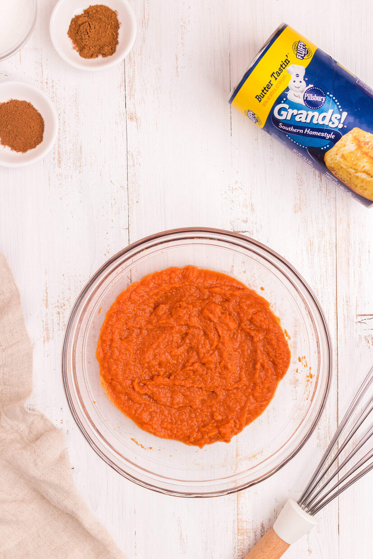 A mixed pumpkin filling in a glass bowl. The mixture is smooth and orange, indicating the ingredients have been fully combined. The same can of biscuits and spice bowls remain nearby.