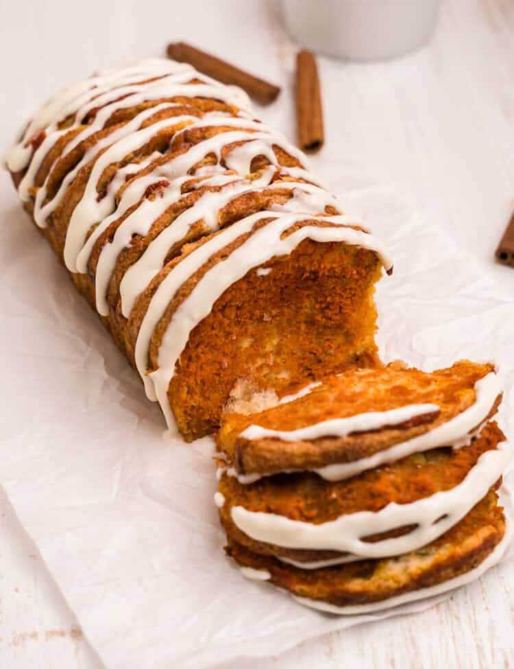 A sliced loaf of pumpkin pull-apart bread with icing on top. A few pieces are pulled forward, revealing the soft, moist pumpkin interior. Cinnamon sticks and a bowl of pumpkin puree are in the background.