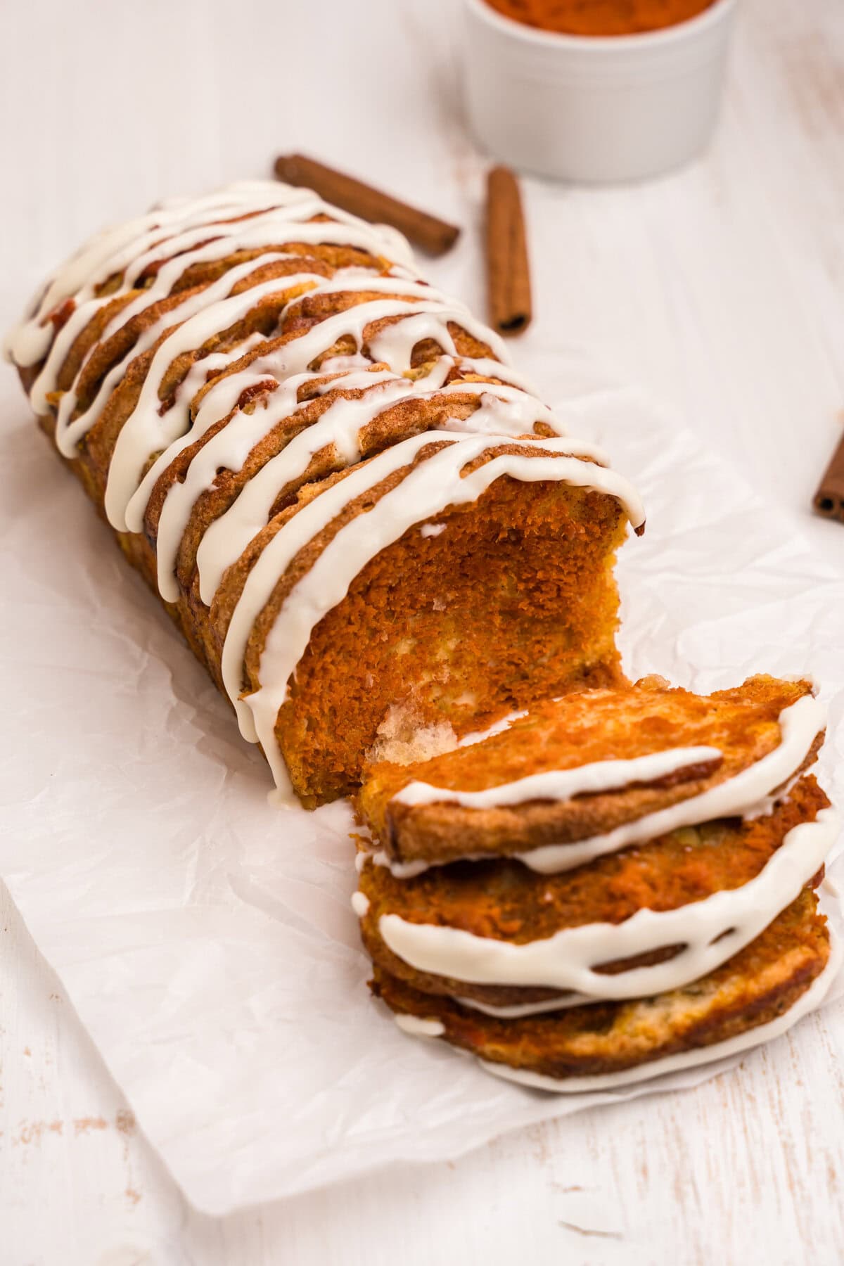 A sliced loaf of pumpkin pull-apart bread with icing on top. A few pieces are pulled forward, revealing the soft, moist pumpkin interior. Cinnamon sticks and a bowl of pumpkin puree are in the background.

