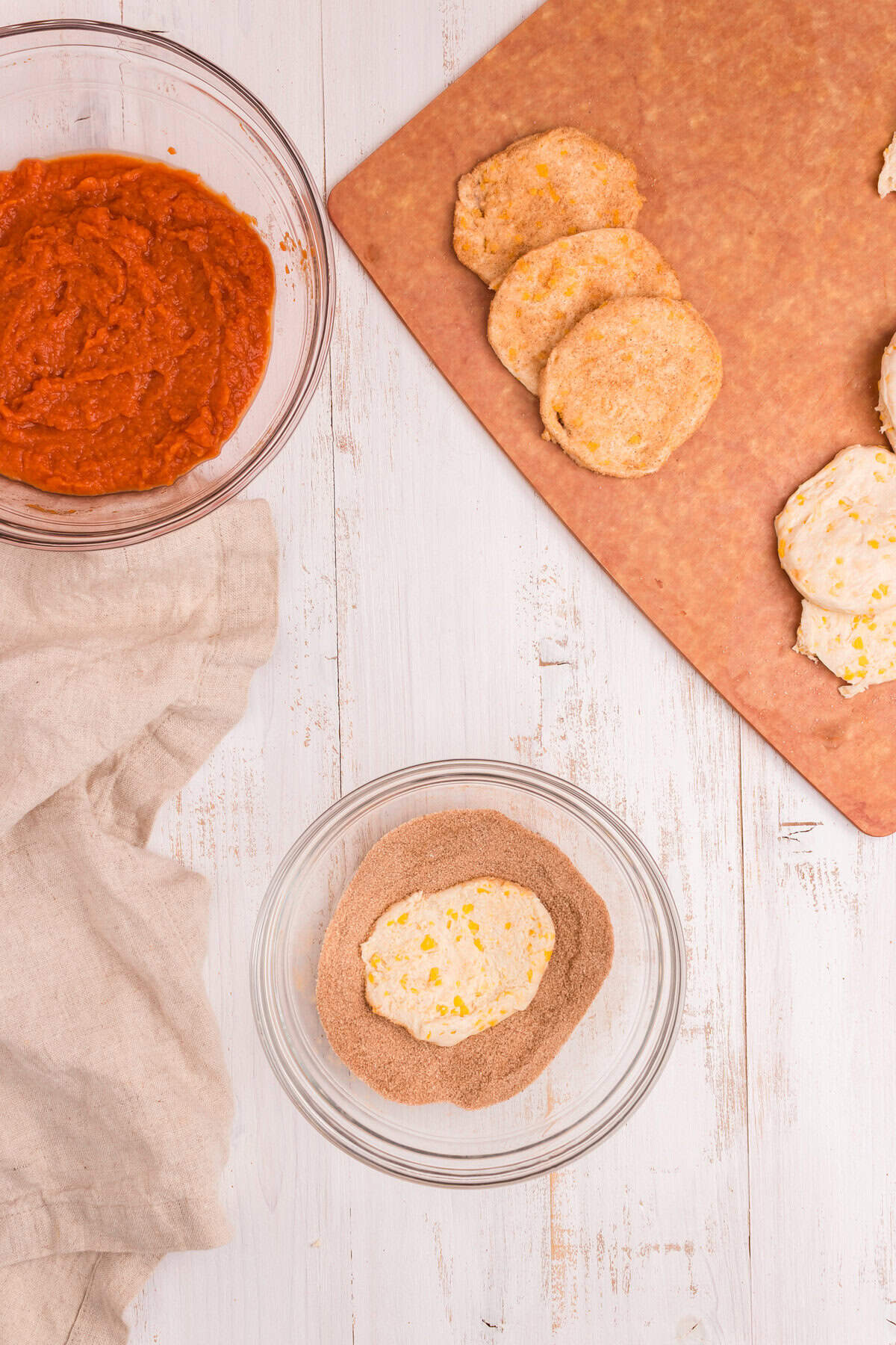 A step further in the process—more biscuit dough rounds are coated in cinnamon sugar and stacked on a cutting board. One dough piece is currently being coated in cinnamon sugar.