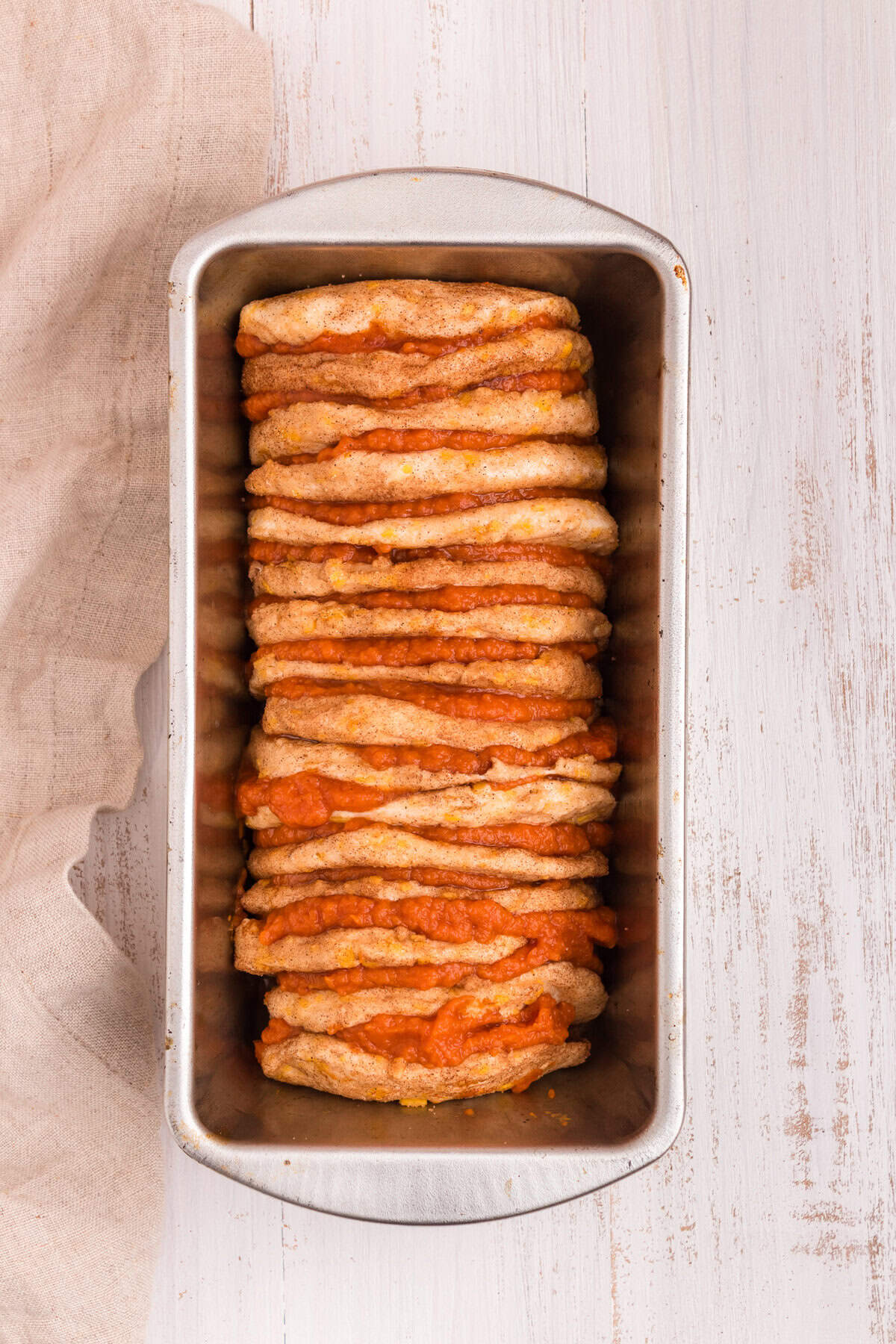 A metal loaf pan filled with stacked layers of cinnamon-sugar-coated biscuit dough, each layered with pumpkin filling. The loaf is ready to be baked.