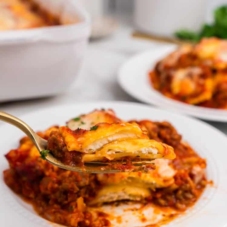 A close-up view of a serving of ravioli lasagna being held up on a fork over a white plate, showing layers of ravioli, meat sauce, and melted cheese.