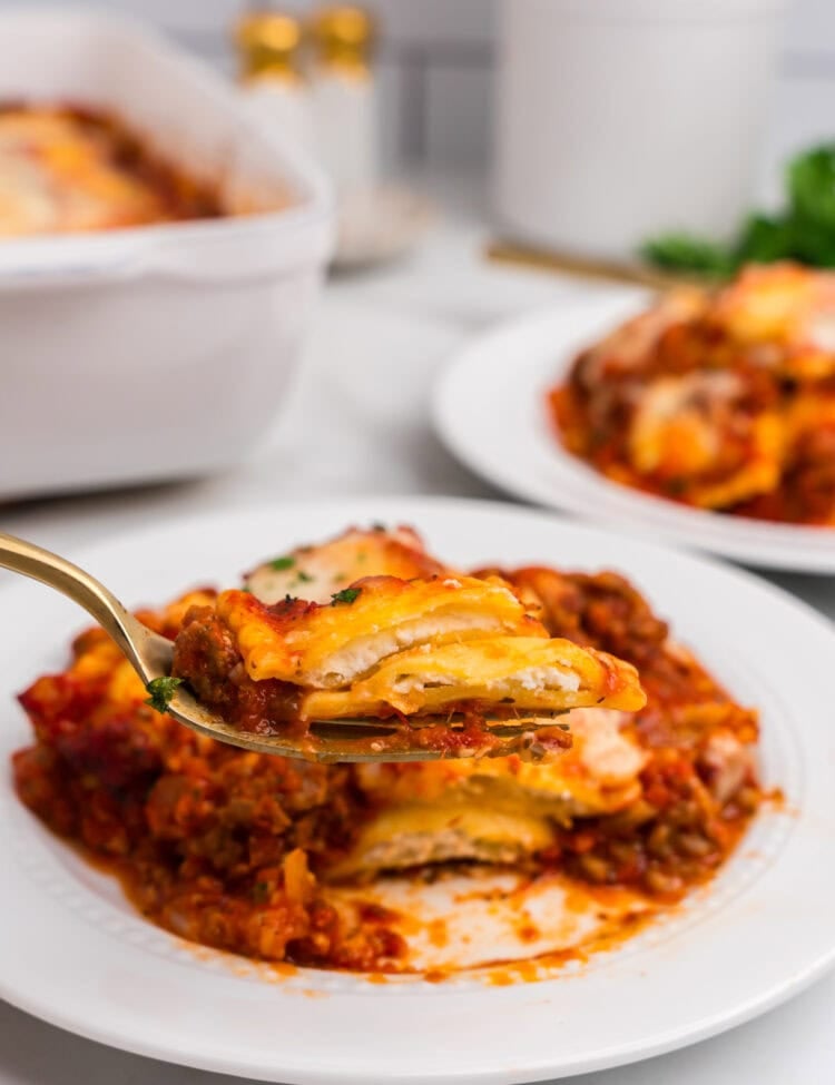 A close-up view of a serving of ravioli lasagna being held up on a fork over a white plate, showing layers of ravioli, meat sauce, and melted cheese.