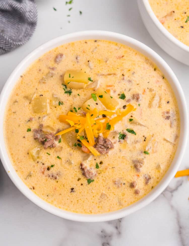 Overhead view of two bowls of creamy cheeseburger soup garnished with shredded cheddar and parsley, with extra shredded cheese on the side.