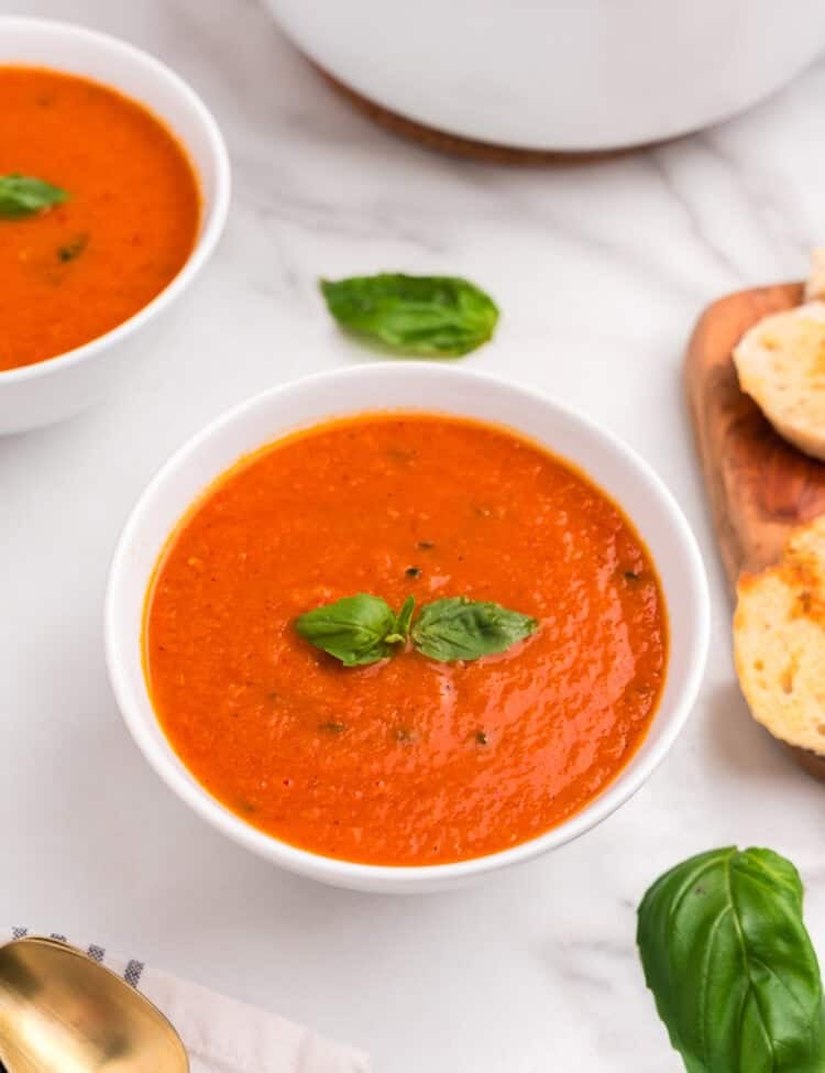 A close-up of a white bowl filled with creamy roasted tomato basil soup, garnished with two fresh basil leaves. The bowl is placed on a marble surface beside a wooden board with toasted bread, loose basil leaves, and a gold spoon on a folded napkin.