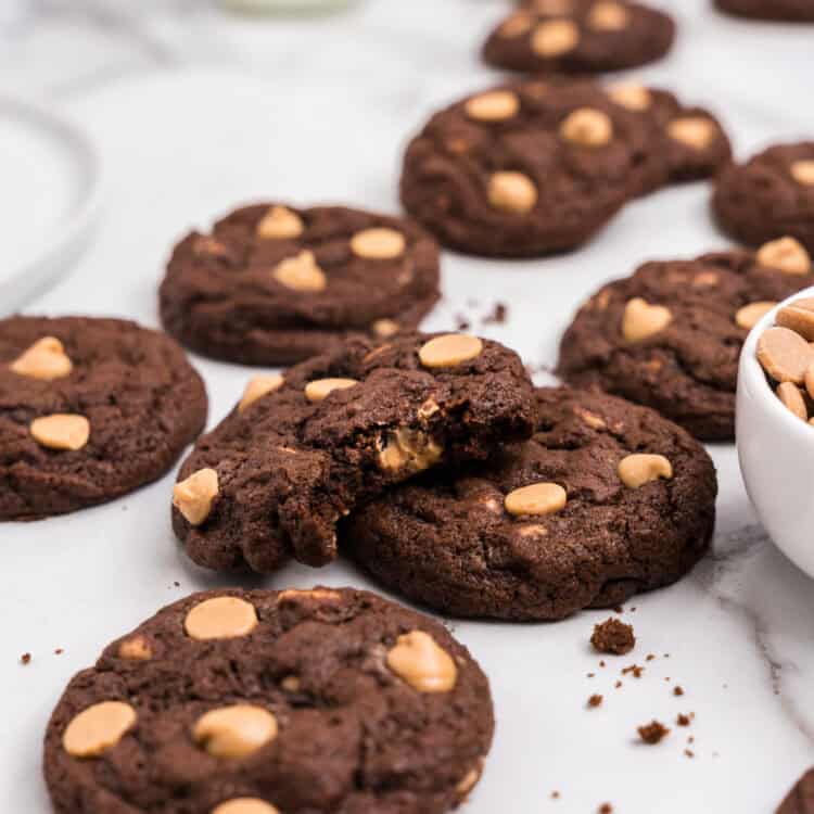 Close-up of a pile of chocolate and peanut butter chip cookies on a white surface, with one cookie broken in half to reveal a gooey interior. A glass of milk and bowl of peanut butter chips are visible in the background.