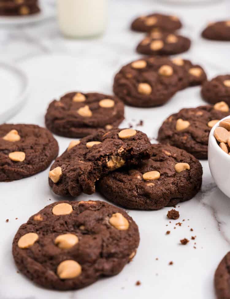 Close-up of a pile of chocolate and peanut butter chip cookies on a white surface, with one cookie broken in half to reveal a gooey interior. A glass of milk and bowl of peanut butter chips are visible in the background.