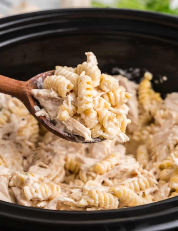 A close-up of a wooden spoon lifting a portion of creamy garlic parmesan chicken pasta from a black slow cooker. The rotini pasta is coated in a rich, cheesy sauce with tender shredded chicken mixed throughout. In the background, the rest of the dish remains in the crockpot, with hints of fresh parsley and garlic visible, adding to the inviting and comforting meal presentation.