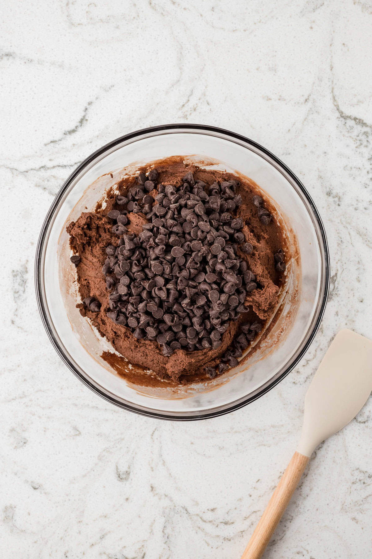 A mixing bowl of chocolate cookie dough with a generous amount of chocolate chips added on top, ready to be folded in.