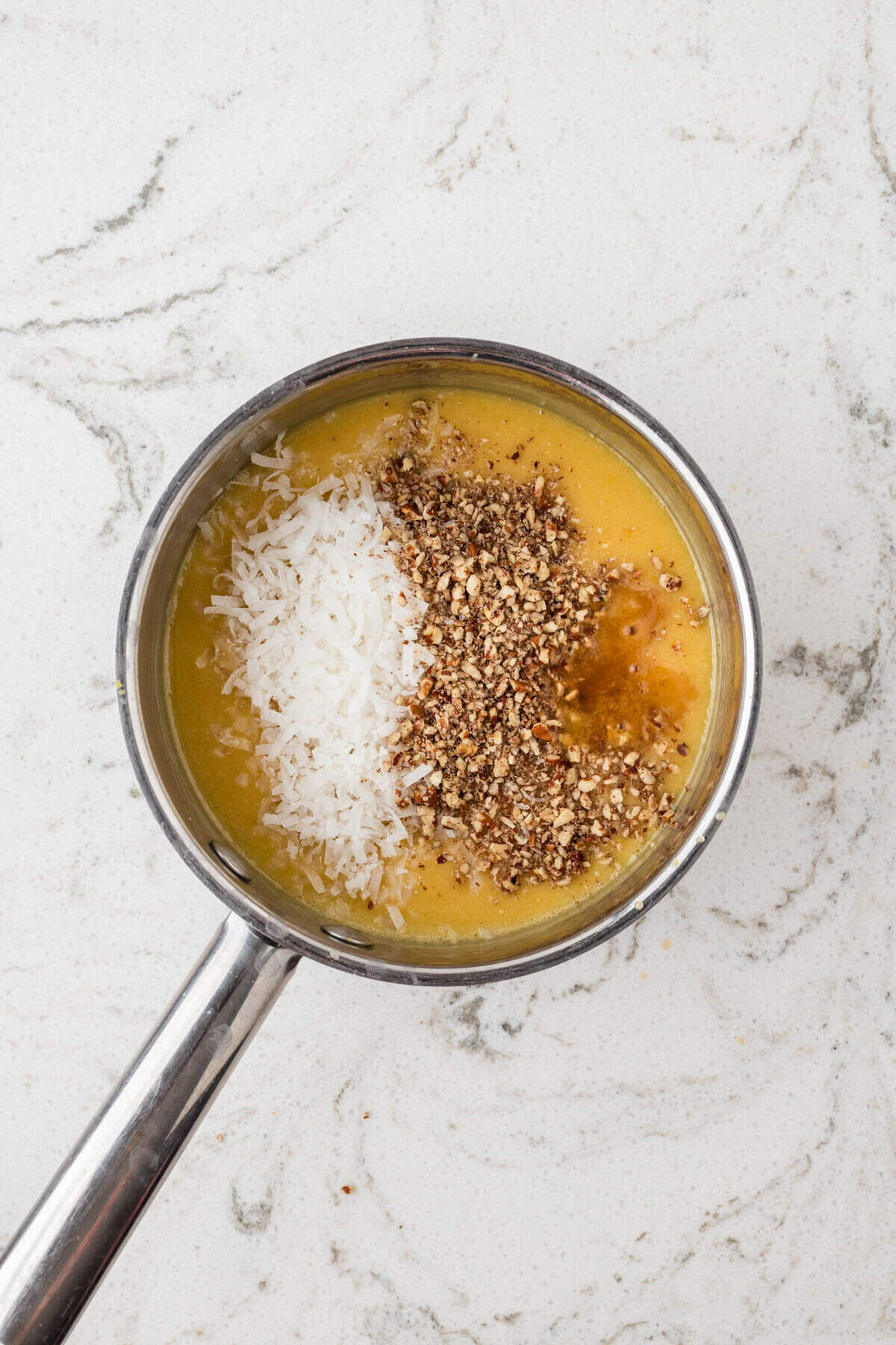 A saucepan with a yellow batter mixture, topped with shredded coconut, chopped pecans, and a splash of vanilla extract, ready to be stirred.