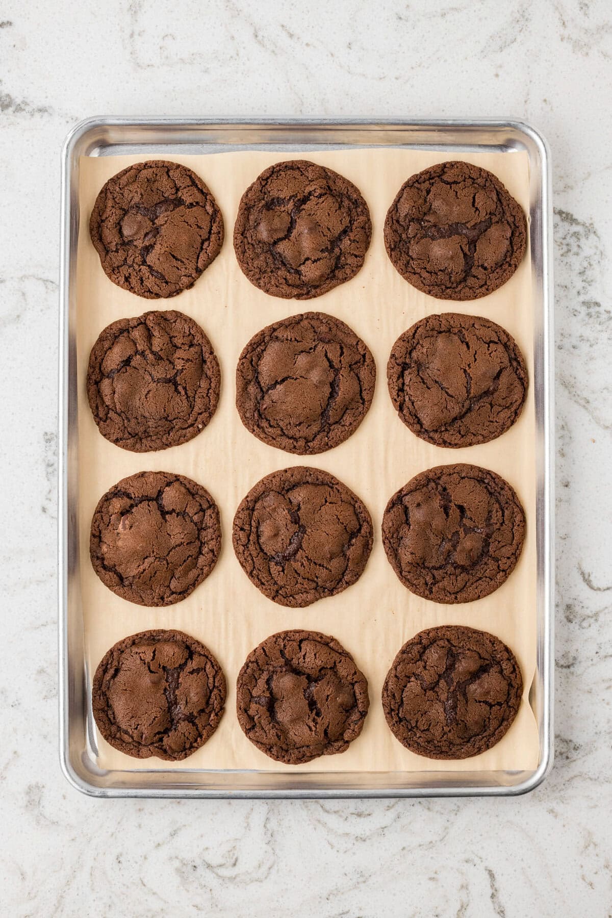A baking tray lined with parchment paper holding twelve freshly baked chocolate cookies with cracked tops.