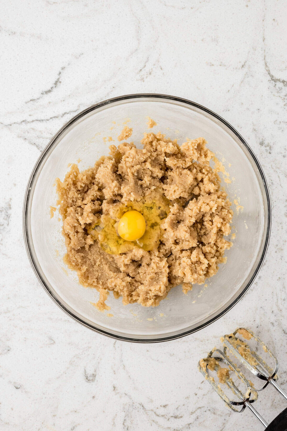 A mixing bowl with a creamed butter and sugar mixture and one cracked egg on top, ready to be blended.