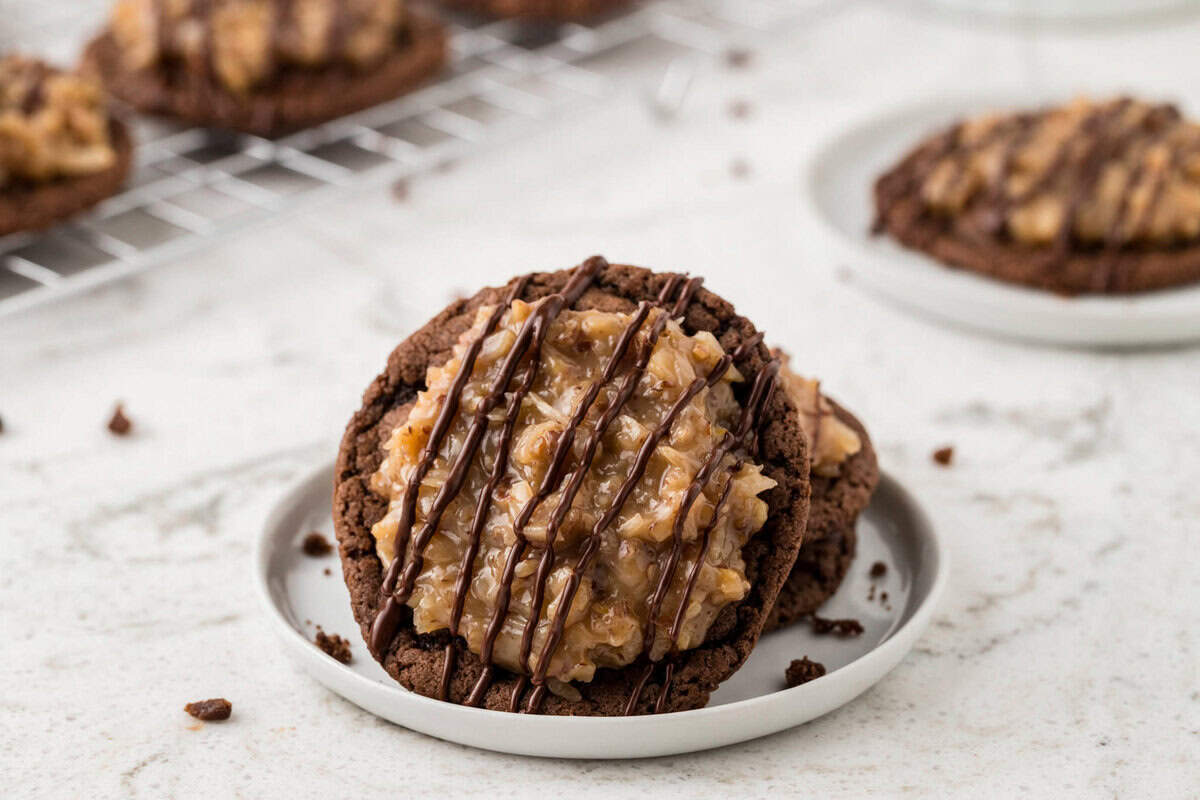 Two German chocolate cookies stacked on a small plate. The cookies have a rich coconut pecan topping and are drizzled with chocolate, with more cookies visible on a cooling rack in the background.